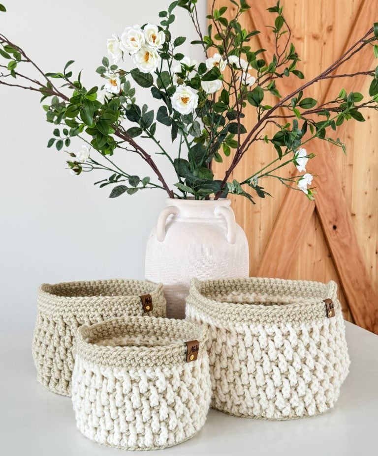 A white vase with white flowers and green leaves stands behind three cream-colored crochet baskets on a white surface.