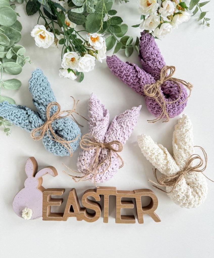 Four crocheted bunny figures in pastel colors, reminiscent of a crochet dishcloth’s texture, are tied with twine and arranged near wooden "EASTER" letters, surrounded by white flowers and green leaves.