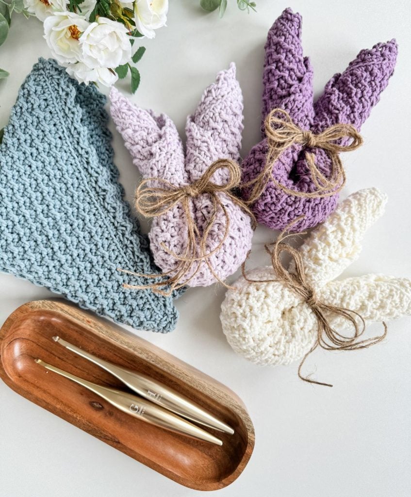 Four knitted bunny-shaped crafts in pastel colors with twine bows are arranged beside crochet hooks, a crochet dishcloth, and white flowers in a wooden tray on a white surface.