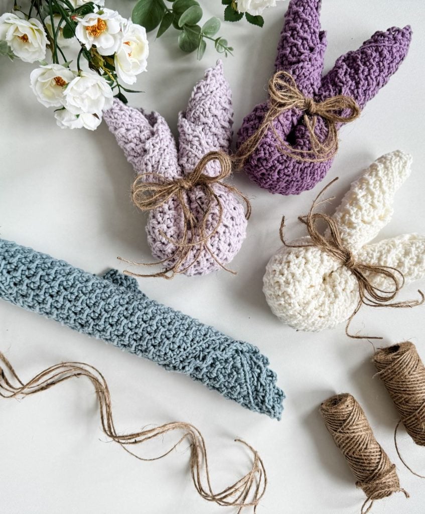 Three knitted bunny-shaped decorations tied with twine, a rolled blue crochet dishcloth, spools of twine, and white flowers arranged on a white surface.
