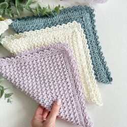 A hand holds a textured, pastel purple crochet dishcloth, with two similar cloths in cream and teal displayed nearby on a white surface beside skeins of matching yarn and green foliage.