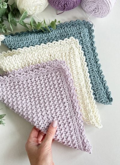 A hand holds a textured, pastel purple crochet dishcloth, with two similar cloths in cream and teal displayed nearby on a white surface beside skeins of matching yarn and green foliage.
