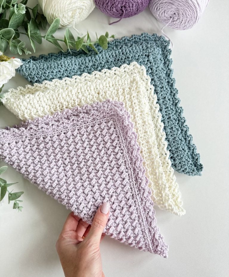 A hand holds a textured, pastel purple crochet dishcloth, with two similar cloths in cream and teal displayed nearby on a white surface beside skeins of matching yarn and green foliage.