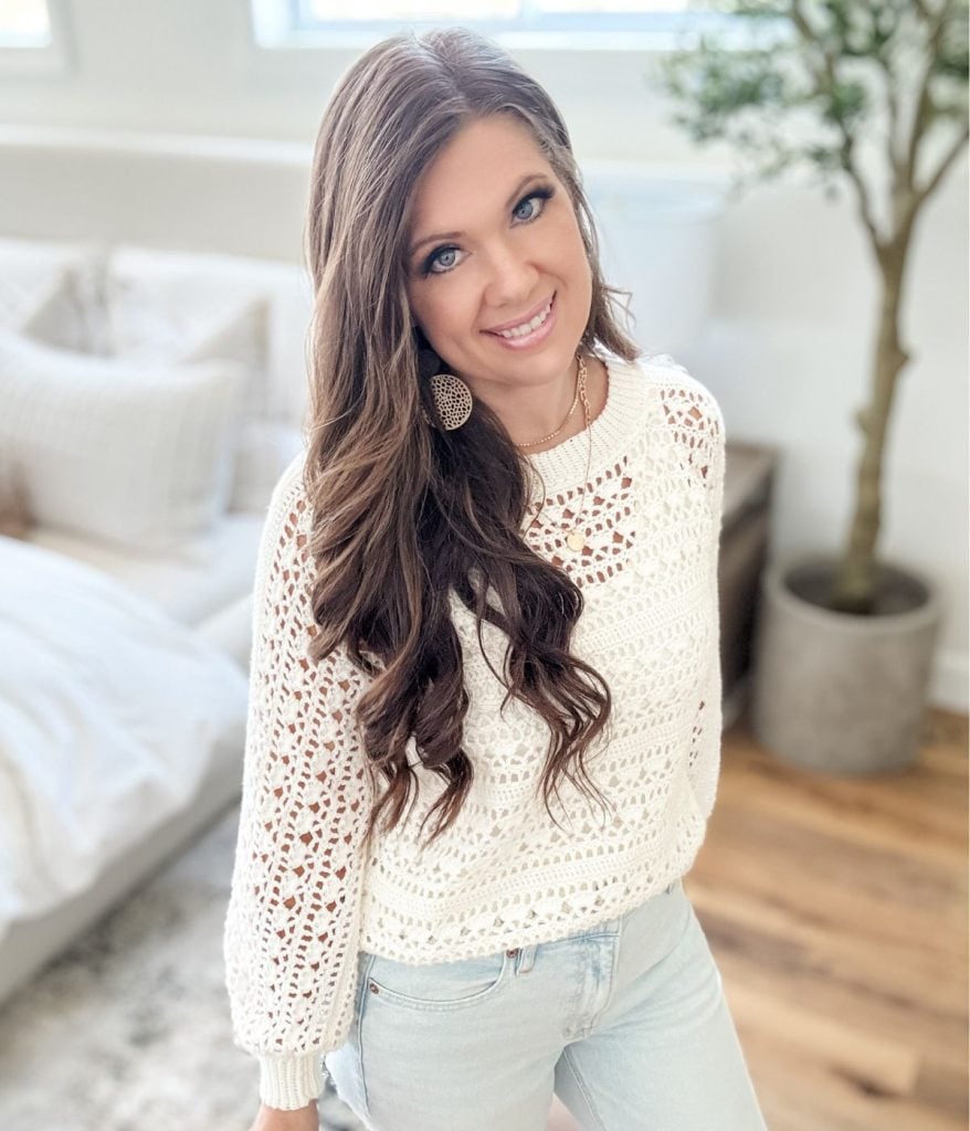 Woman with long brown hair wearing a Caribbean Sea Sweater and light blue jeans standing in a bright, modern room with a plant in the background.