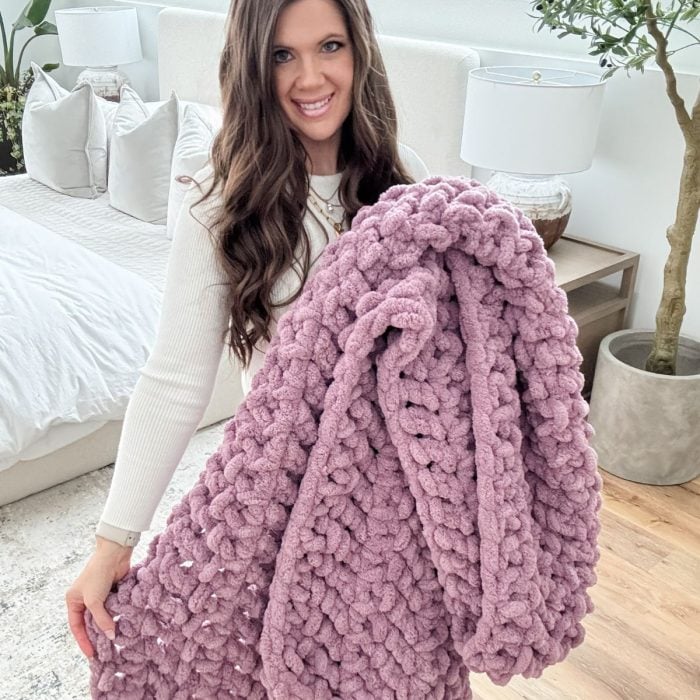 A woman standing in a bright bedroom holds a chunky, knitted mauve jumbo blanket and smiles at the camera.