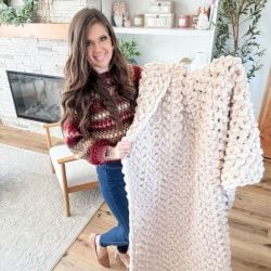 A woman stands in a living room holding a chunky, light-colored jumbo blanket, with yarn and decor visible on shelves in the background.