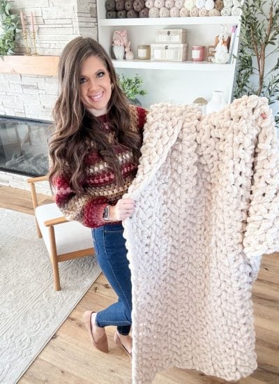 A woman stands in a living room holding a chunky, light-colored jumbo blanket, with yarn and decor visible on shelves in the background.