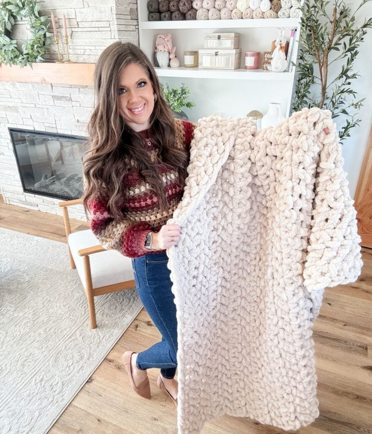 A woman stands in a living room holding a chunky, light-colored jumbo blanket, with yarn and decor visible on shelves in the background.