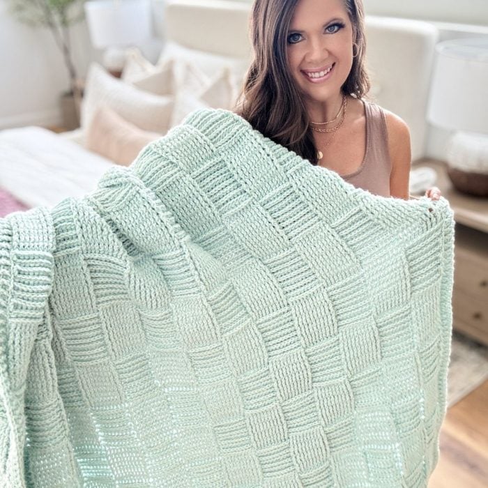 A woman stands in a bedroom holding up a large, light blue Basket Weave Blanket with a beautiful checkered pattern.