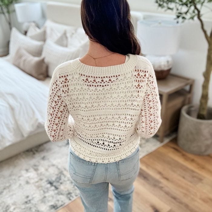 A woman with long brown hair stands indoors facing away, wearing a white Caribbean Sea Sweater and light blue jeans. A bed, nightstand, lamp, and potted plant are visible in the background.