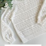 A cream-colored Caribbean Sea Sweater with intricate lace crochet patterns is displayed on a flat surface beside green foliage and white flowers. Text reads "Caribbean Sea Sweater Crochet Pattern & Video.