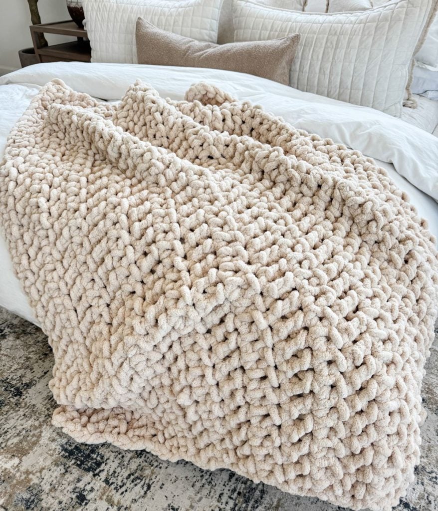 A chunky, beige jumbo blanket is draped over the edge of a bed, with white bedding and neutral pillows in the background.