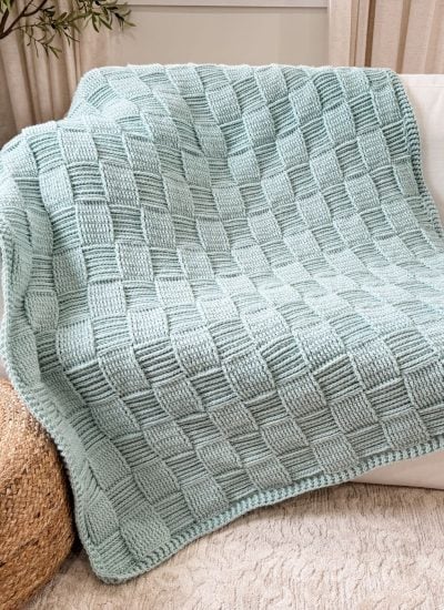 A light blue Basket Weave Blanket with a checkered textured pattern is draped over a white sofa next to a woven basket.