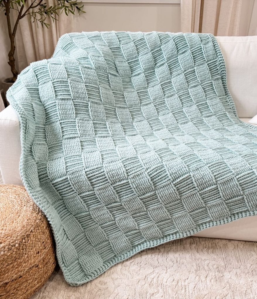 A light blue Basket Weave Blanket with a checkered textured pattern is draped over a white sofa next to a woven basket.
