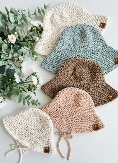 Five crochet sunhats in white, light blue, brown, beige, and cream are arranged in a row next to white flowers and green leaves on a light surface.