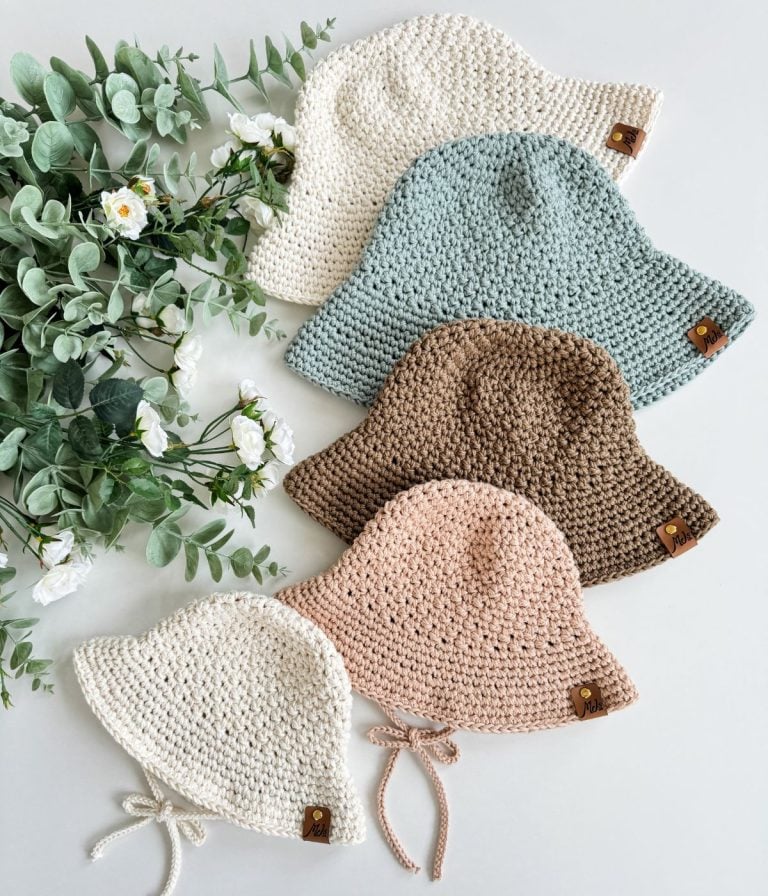 Five crochet sunhats in white, light blue, brown, beige, and cream are arranged in a row next to white flowers and green leaves on a light surface.