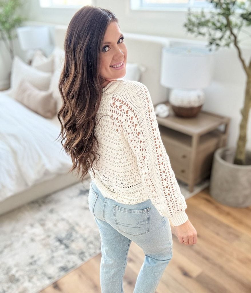 Woman with long brown hair wearing a Caribbean Sea Sweater and light blue jeans stands in a bright bedroom, looking over her shoulder and smiling at the camera.