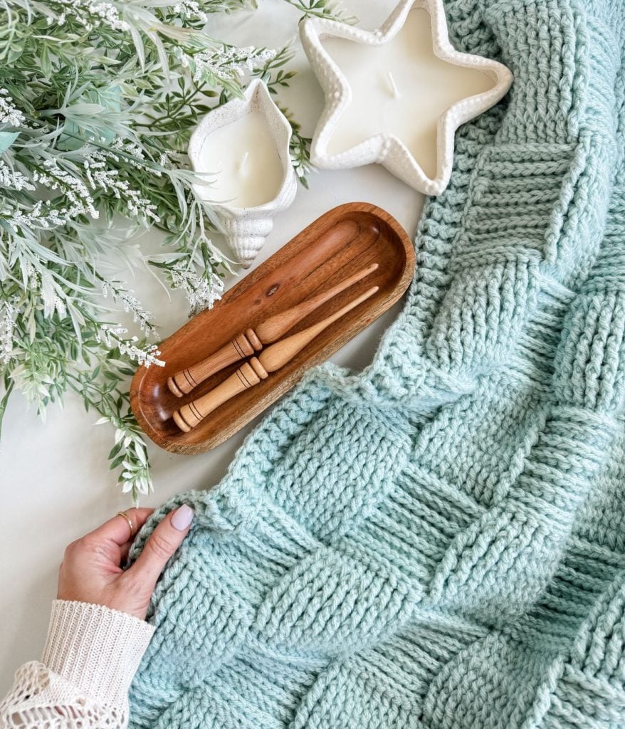A hand holds a light blue Basket Weave Blanket next to a wooden tray with crochet hooks, a star-shaped candle, a shell-shaped candle, and green leafy branches.