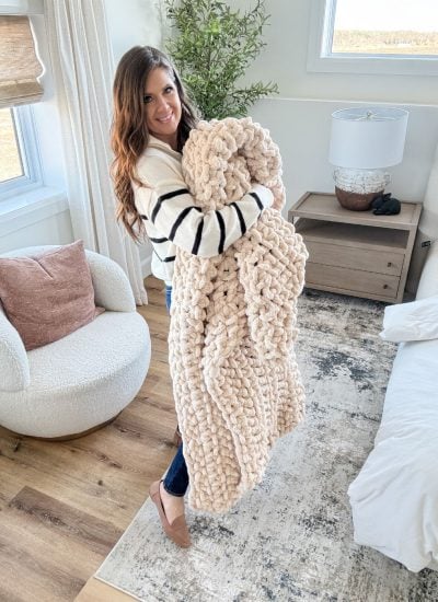 A woman stands in a bedroom holding a jumbo blanket, next to a white chair and a wooden nightstand with a lamp and decor.