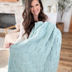 A woman with long brown hair holds up a light blue Basket Weave Blanket while smiling in a cozy living room.