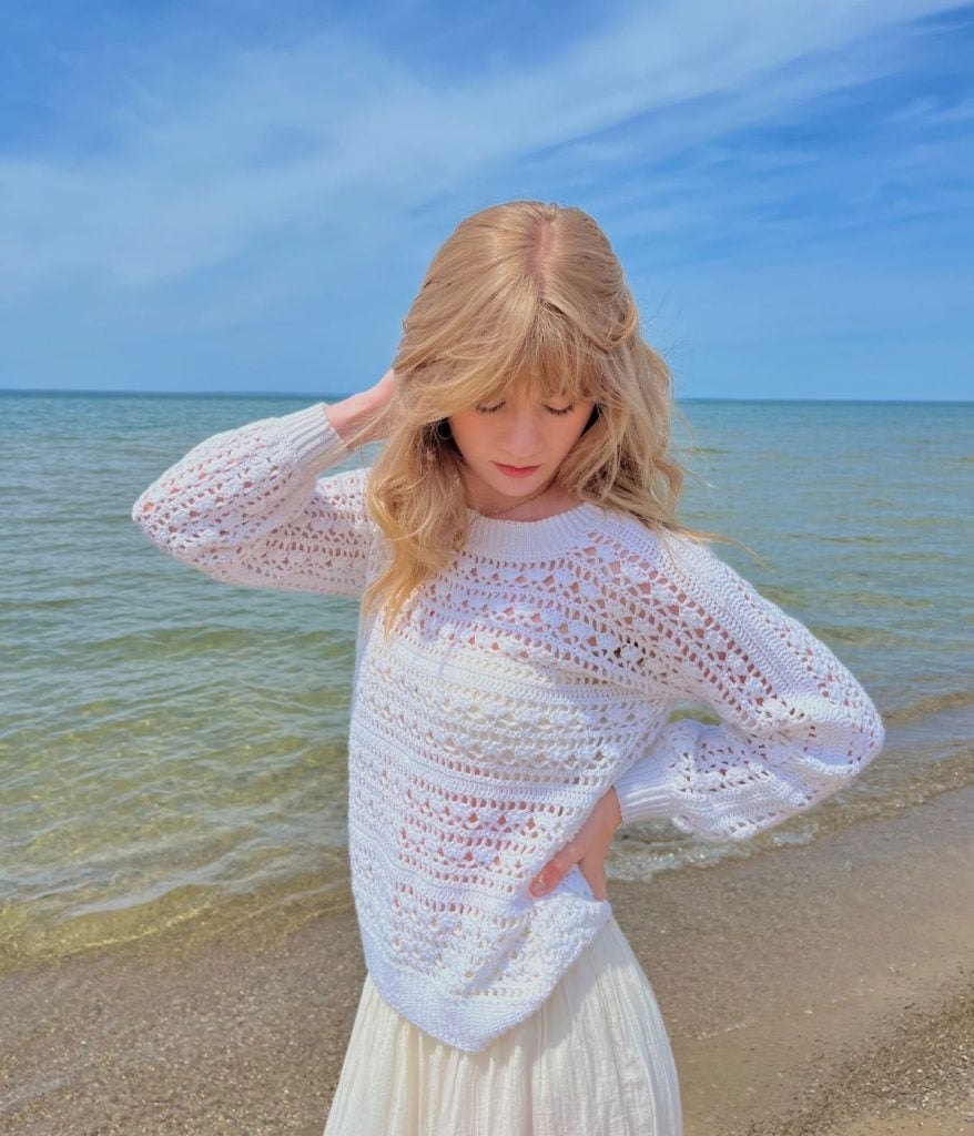 A person with blonde hair wearing a white knit Caribbean Sea sweater stands on a sandy beach near the water under a blue sky.