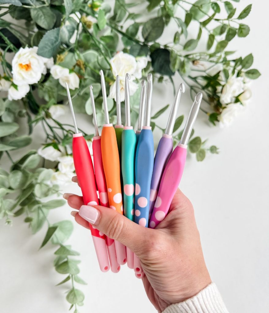 A hand holds eight colorful crochet hooks with soft handles—perfect for crafting your next Caribbean Sea Sweater—in front of white flowers and green leaves.