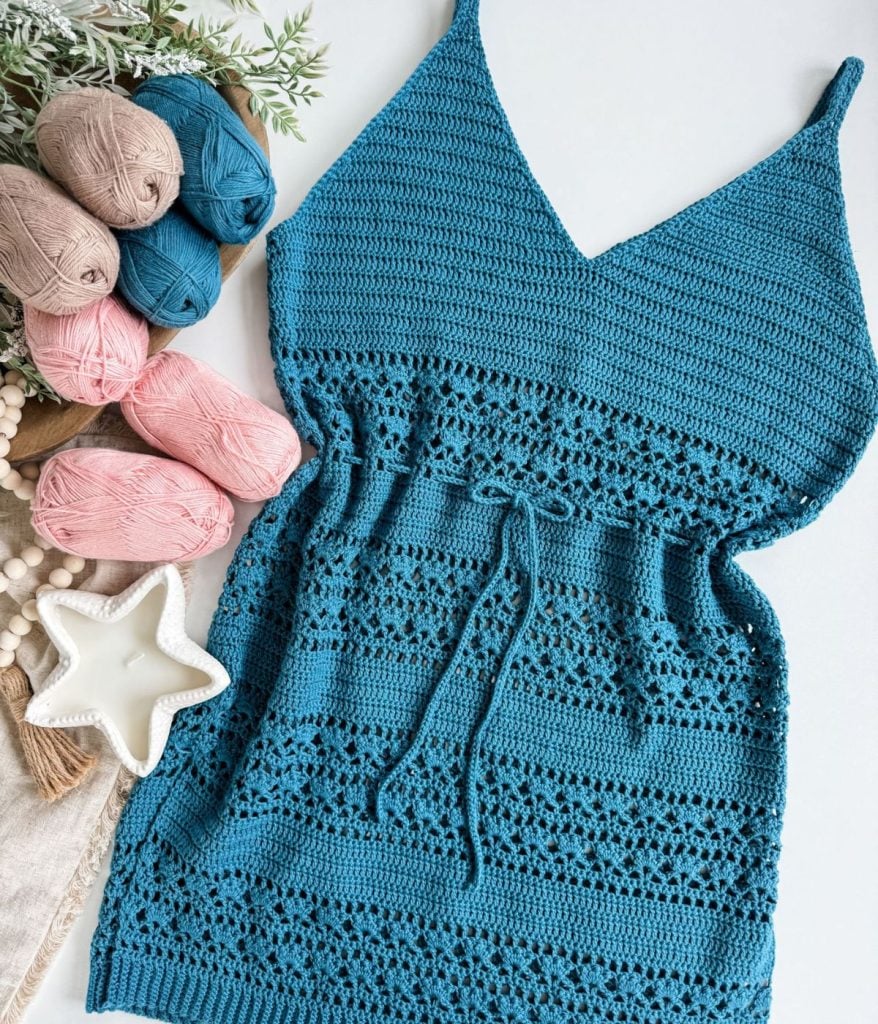 A crocheted teal dress with lace detailing is displayed next to skeins of yarn in blue, pink, and beige—the perfect palette for crafting a Caribbean Sea Sweater—alongside a ceramic star-shaped dish and some greenery.