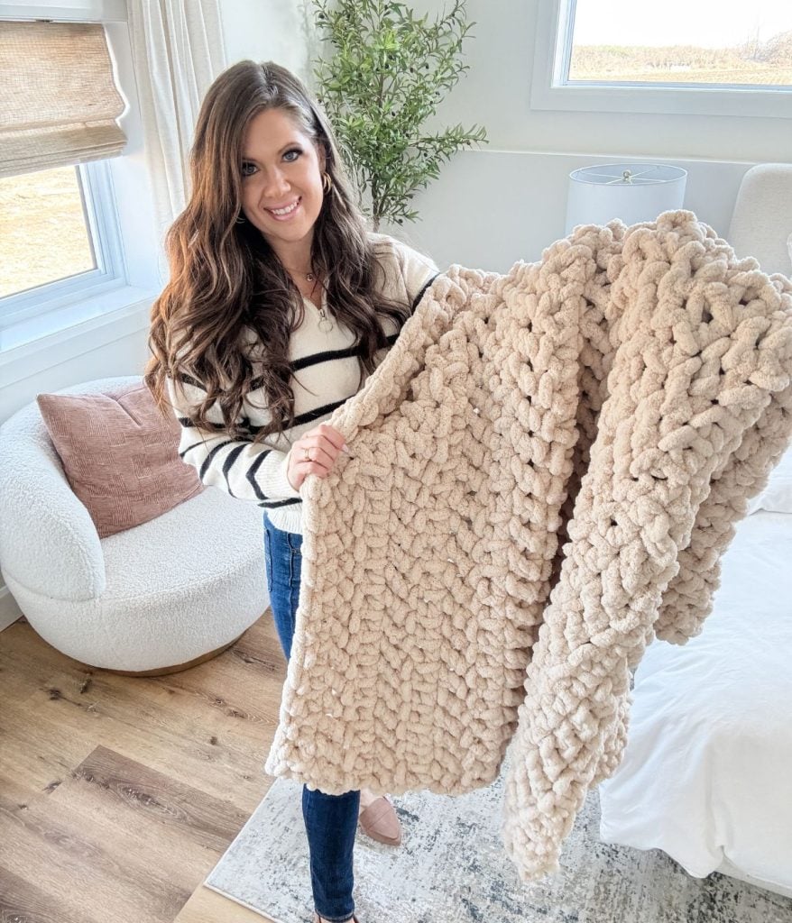 A woman indoors holds up a large, chunky-knit jumbo blanket, standing next to a white chair and a green plant.