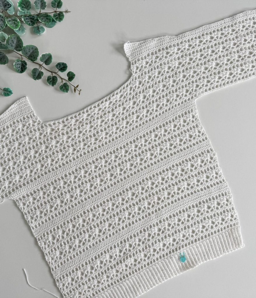 White crocheted Caribbean Sea Sweater with a lacy, textured pattern is laid flat on a light surface, with green eucalyptus leaves visible in the top left corner.