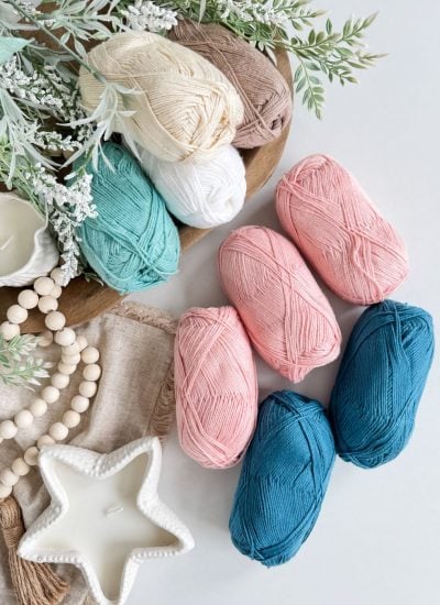 Balls of yarn in pastel pink, teal, beige, white, and dark blue—perfect for a Caribbean Sea Sweater—are arranged on a table with candles, wooden beads, fabric, and green foliage.