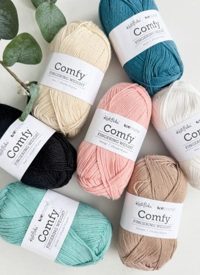 Six skeins of Comfy Fingering Weight yarn in black, cream, white, teal, peach, and tan are arranged on a white surface next to a green leafy branch—perfect for crafting your own Caribbean Sea Sweater.