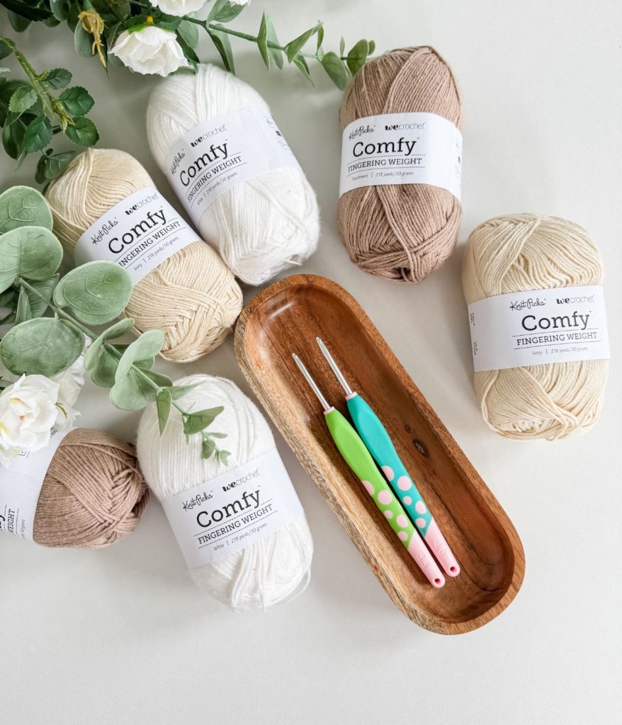 Four skeins of neutral-colored yarn, two crochet hooks, and a wooden tray are arranged on a white surface with artificial greenery and white flowers—perfect for starting your Caribbean Sea Sweater project.