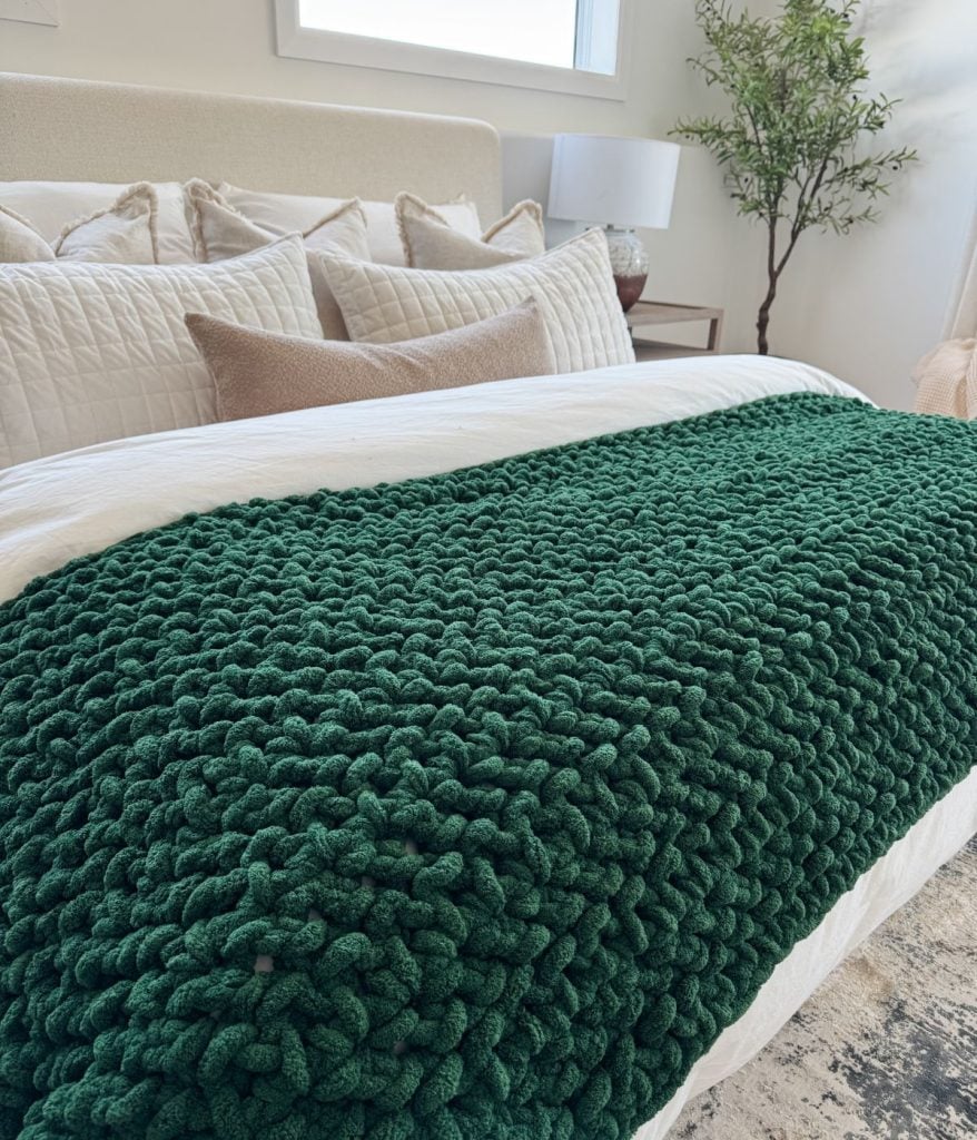 A neatly made bed with neutral pillows and bedding, featuring a chunky dark green jumbo blanket at the foot; a lamp and potted plant add a cozy touch in the background.