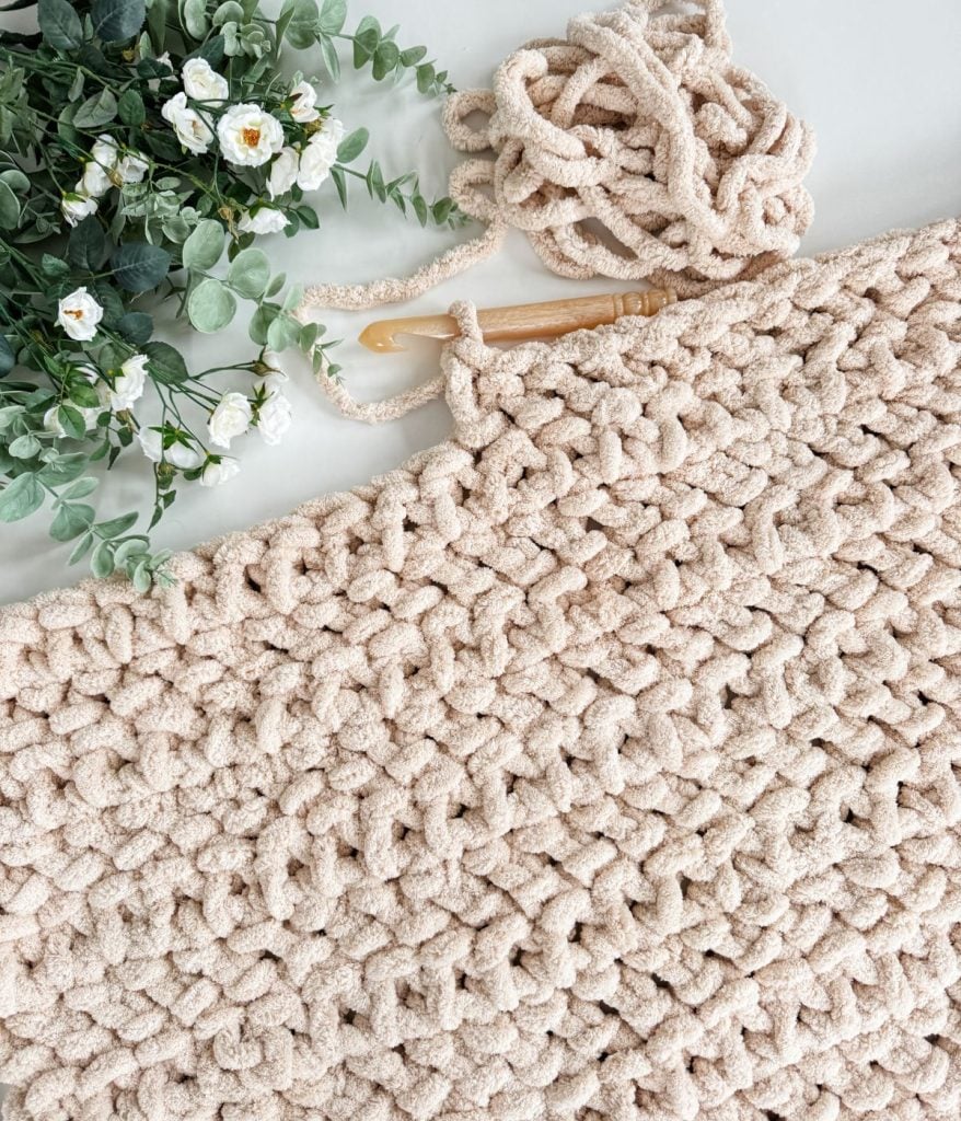 A partially completed jumbo blanket with a crochet hook and yarn sits beside a green and white floral arrangement.