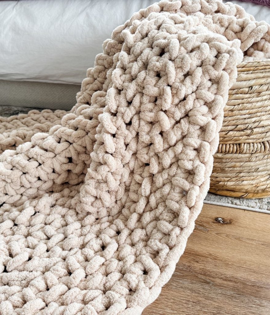 A thick, chunky knit jumbo blanket in beige is draped over the edge of a woven basket on a wood floor, near a bed.