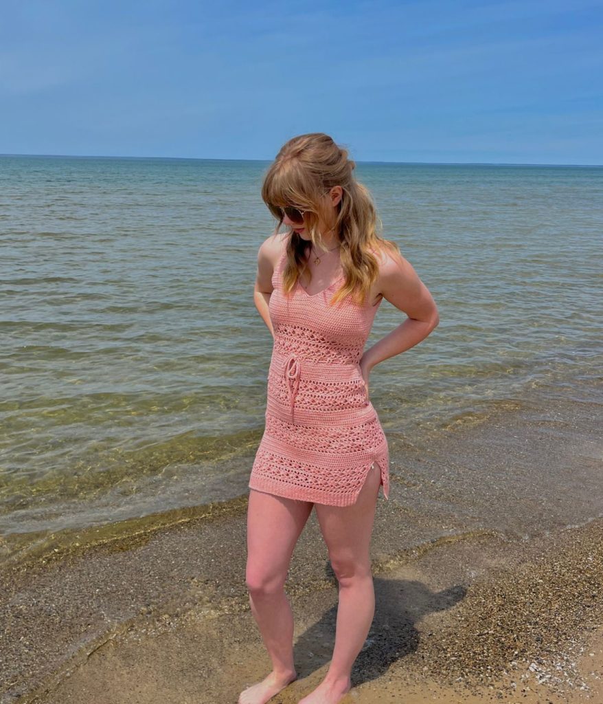 A woman in a pink crochet dress stands barefoot on a sandy beach, the Caribbean Sea sweatered in blue behind her under the clear sky.