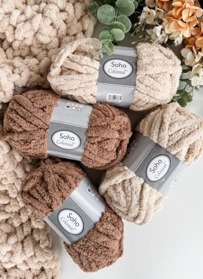Four skeins of chunky Soho Colossal yarn in beige and brown shades are arranged on a white surface, perfect for creating a cozy jumbo blanket. Greenery and artificial flowers accent the background.