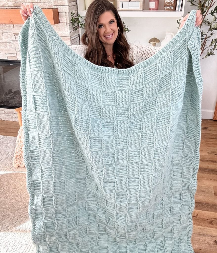 A woman indoors holds up a large light blue Basket Weave Blanket featuring a stylish checkered square pattern.