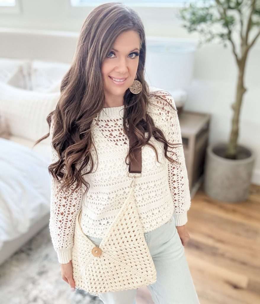 Woman with long brown hair wears a white Caribbean Sea Sweater and holds a matching crochet bag. She stands indoors with light-colored decor and a plant in the background.