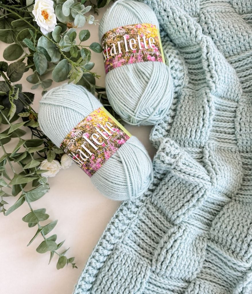 Two skeins of mint green yarn labeled "Starlette" rest beside a partially knitted basket weave blanket and some artificial greenery.