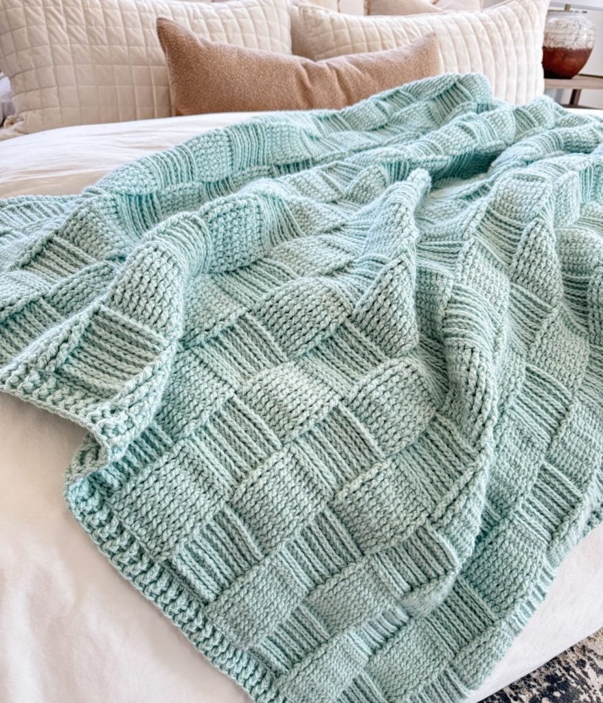 A light blue Basket Weave Blanket with a checkered pattern is spread out on a bed with cream and tan pillows.
