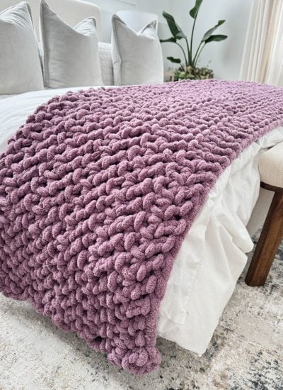 A jumbo blanket in chunky knit purple is draped over the end of a white bed, complemented by neutral-toned pillows, a bench, and a potted plant in the background.