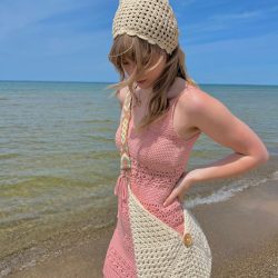 A woman in a pink crochet dress and matching hat stands on a sandy beach with a woven bag, gazing down near the Caribbean Sea Sweater-blue water under a clear sky.