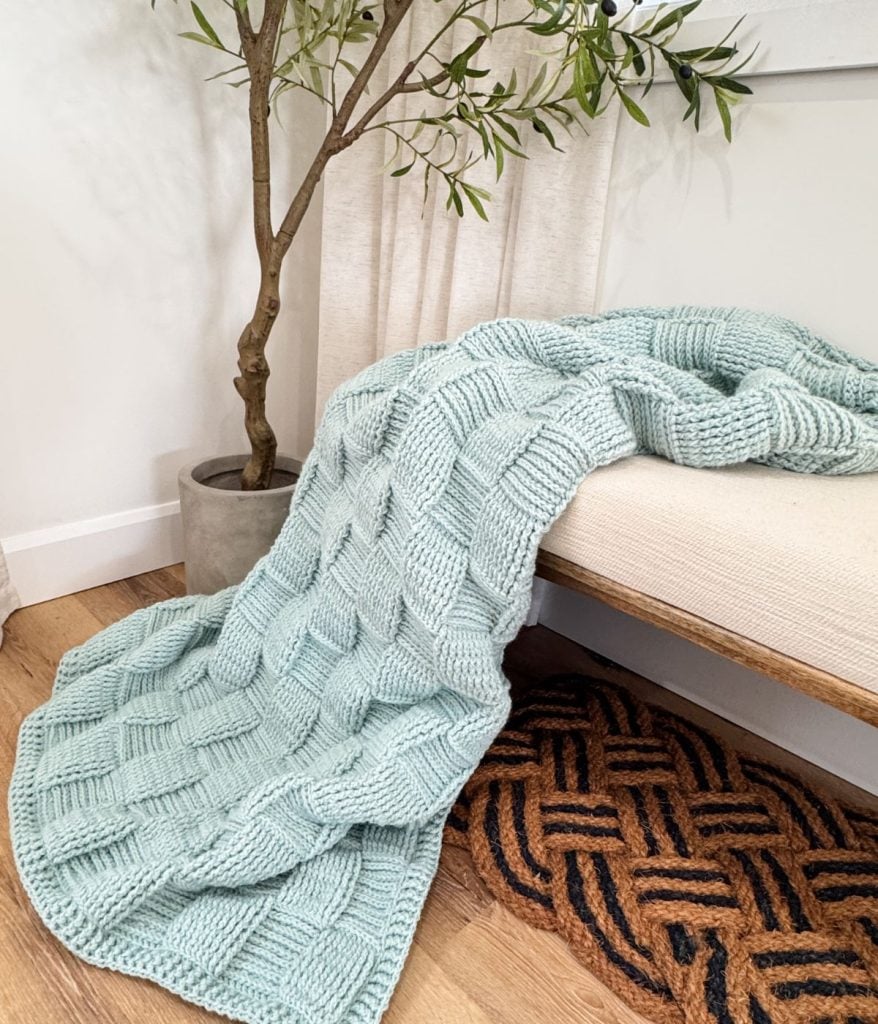 A light blue, chunky Basket Weave Blanket with a checkered pattern is draped over a beige bench beside a potted plant and a brown woven rug.