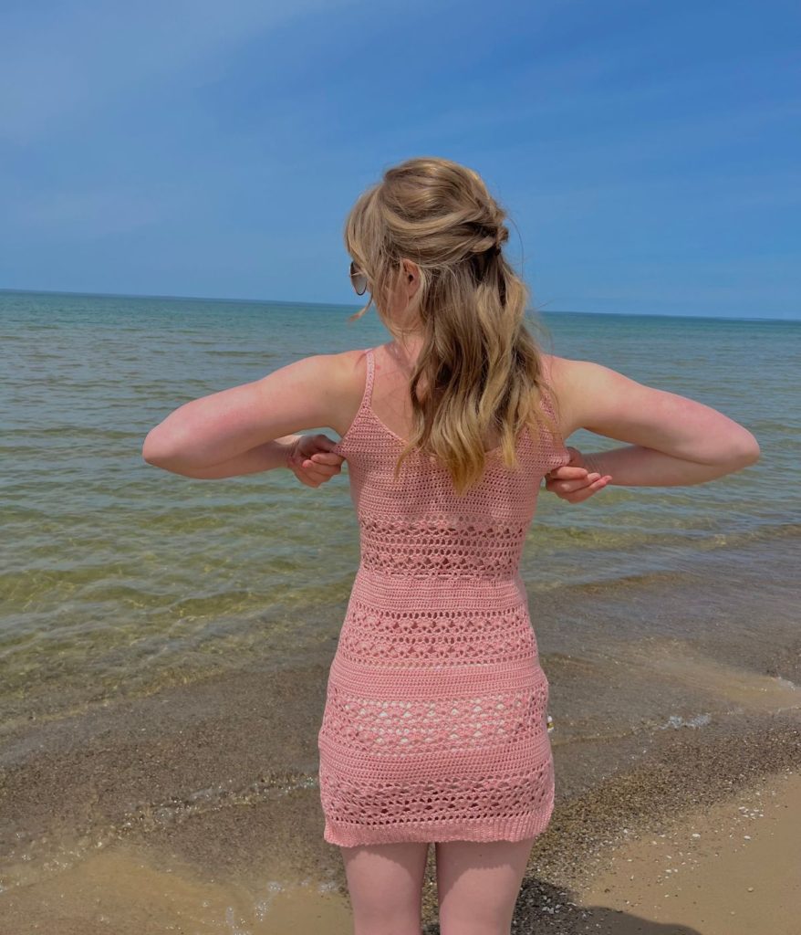 A woman with light brown hair in a pink crochet dress stands facing the lake, adjusting her straps on a sandy beach under a clear sky, her style echoing the breezy charm of a Caribbean Sea Sweater.