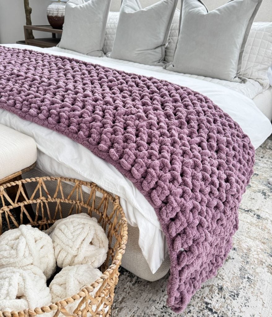 A chunky purple jumbo blanket is draped over the foot of a bed with white pillows, next to a woven basket holding white yarn balls.
