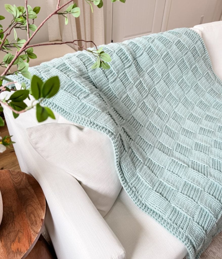 A light blue Basket Weave Blanket with a textured square pattern is draped over a white sofa, with part of a green plant visible in the foreground.