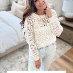 Woman stands in a bright bedroom wearing the Caribbean Sea Sweater—a white crocheted design paired with light jeans. Text below reads: "Caribbean Sea Sweater Crochet Pattern & Video.