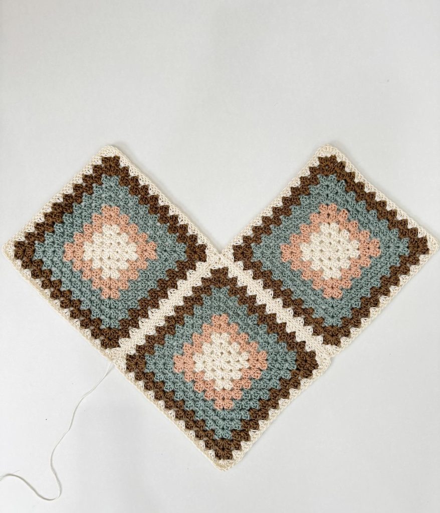 Three crocheted granny squares in blue, pink, brown, and white are joined together in a V shape on a white background, perfect for creating a colorful Granny Square Bag or adding flair to your next Crochet Pattern.
