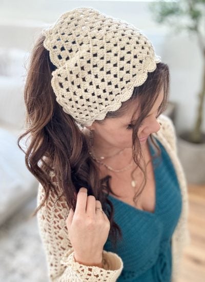 Woman with long brown hair wears a stylish crochet bandana as a cream headscarf and a blue top, standing indoors with neutral décor in the background.